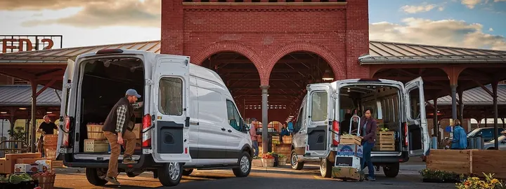 Two Ford Transit cargo vans with their rear doors open, being loaded at a city market