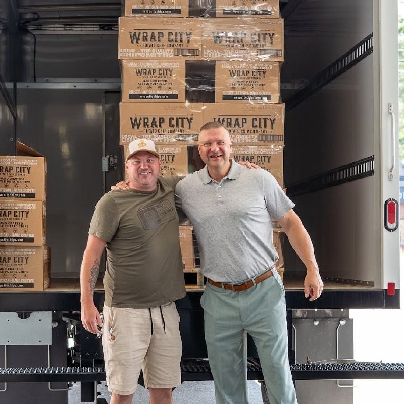 Two men embracing in front of stacked Wrap City Chips boxes